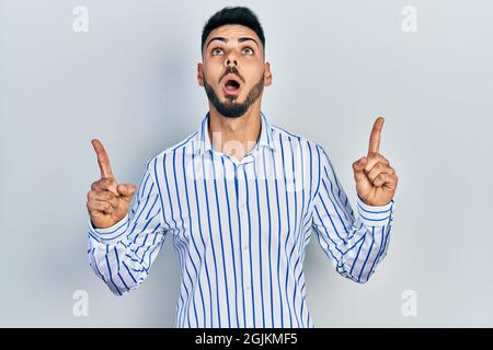 Giovane ispanico con barba indossando una maglietta casual a righe stupito e sorpreso guardando in alto e puntando con le dita e le braccia sollevate. Foto Stock