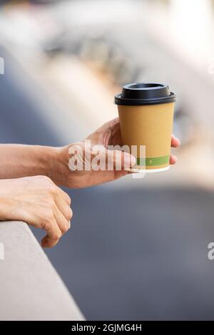 Mani che tengono una tazza di carta da caffè monouso. Giovane donna in un balcone bere caffè da tazza usa e getta con una vista offuscata della città. Foto Stock