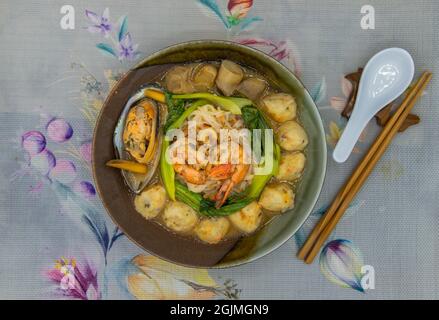 Minestra di spaghetti di riso sottili con cozze, gamberi, polpette, funghi e verdure serviti in un recipiente in ceramica con bacchette e cucchiaio. Stile asiatico per Foto Stock