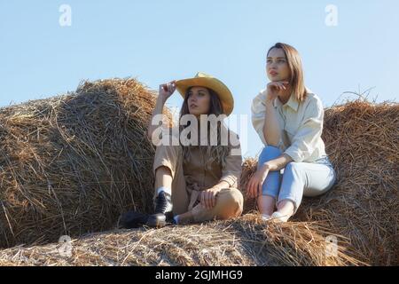 Due giovani donne bruna e bionda sono sedute sui rotoli di fieno. Romantiche ragazze di campagna sul fieno. Foto Stock