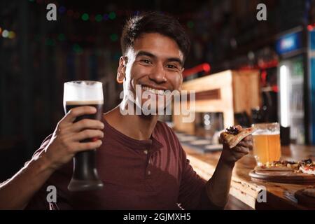 Un uomo allegro e bello sorridendo gioiosamente, reggendo una fetta di pizza e un bicchiere di birra Foto Stock