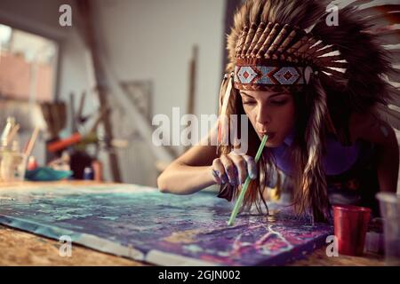 Una giovane artista femminile con un cofano di guerra sta usando una tecnica di paglia mentre lavora ad una nuova pittura in un'atmosfera creativa nello studio. Arte, pittori Foto Stock