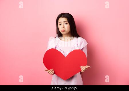 Concetto di San Valentino. Single teenage asian girl vuole innamorarsi, guardando triste e solitario alla macchina fotografica, sulking afflitto in festa degli amanti, tenendo grande Foto Stock