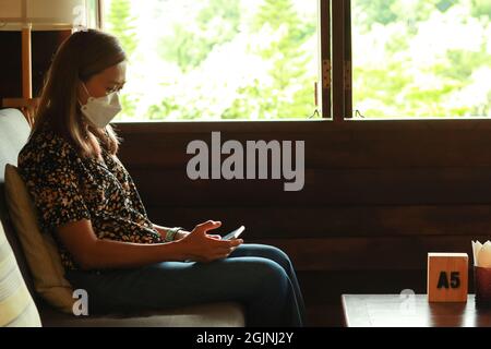 Donna asiatica che indossa maschera viso seduta nella stanza vicino alla finestra e guardando lo smartphone in mani. Foto in interni con ritratto retroilluminato. Foto Stock