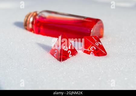 Immagine ravvicinata di due dadi di gioco di ruolo a 4 lati rossi sulla neve. Sullo sfondo una bottiglia di vetro con tappo e liquido rosso Foto Stock