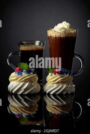 Piccoli dessert Pavlova con lampone, mirtillo e menta. Torte, caffè nero e cioccolata calda con panna montata. Sfondo nero riflettente. Foto Stock