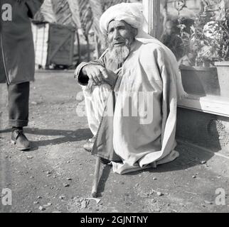 1950, storico, locale, afghano maschio con una barba, headdress e coperta su di lui seduto, squattando in una strada vicino a Kabul, Afghanistan. Foto Stock