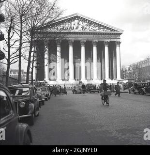 1950, vista storica della chiesa cattolica, la Madeleine con automobili dell'epoca parcheggiate sulla strada acciottolata e due maschi a consegna biciclette con cesti frontali, Parigi, Francia. Costruito come un tempio romano con le sue grandi colonne a testimonianza della gloria militare di Napoleone, fu poi chiamato dal compagno di Gesù, Maria Maddalena. Foto Stock