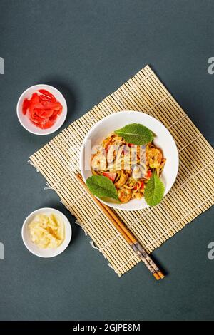 Noodles WOK con frutti di mare e verdure. Cozze, gamberetti e calamari fritti in olio. Delizioso pranzo asiatico in una ciotola bianca su un tappeto di bambù. Mescolare il noo friggere Foto Stock