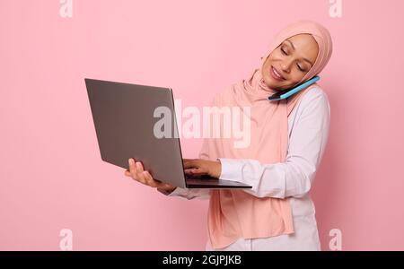 Concetto di una donna d'affari araba musulmana impegnata, freelancer in hijab, parlare su un telefono cellulare mentre si lavora su un portatile allo stesso tempo. Isolato su p Foto Stock