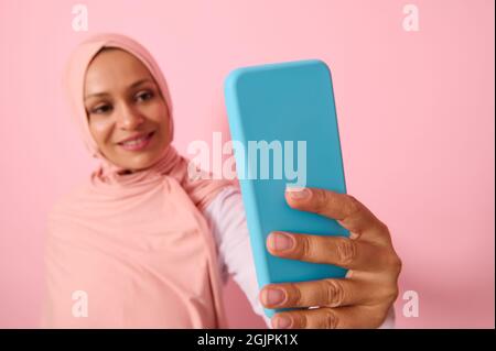 Morbido fuoco sullo smartphone in blu cover in braccia distese di una donna musulmana araba indossando abiti islamici religiosi tradizionali, hijab rosa, e smil Foto Stock