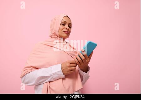 Donna musulmana matura di etnia mediorientale in hijab rosa con smartphone in copertina blu sulle mani, isolata su sfondo rosa pastello con copia Foto Stock