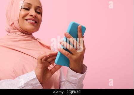 Sorridendo con un sorriso toothy donna musulmana matura di etnia mediorientale in hijab rosa con smartphone in copertura blu sulle sue mani, isolato su p rosa Foto Stock