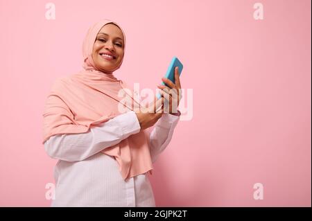 Sorridendo con il sorriso toothy donna musulmana matura di etnia di Medio Oriente in hijab colorato con smartphone in copertura blu sulle mani, guarda la macchina fotografica, è Foto Stock