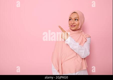 Splendida donna musulmana araba con testa coperta in hijab guardando lo sfondo rosa e puntando il suo indice dito che indica lo spazio di copia per il testo. Foto Stock