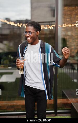 Ritratto verticale del giovane afro-americano che tiene un drink mentre si gode una festa all'aperto sul tetto Foto Stock