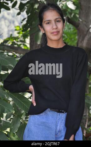 Ritratto di una giovane ragazza affascinante in piedi fuori e indossando una felpa nera e azzurro jean e posando con la mano sui fianchi e guardando la macchina fotografica Foto Stock