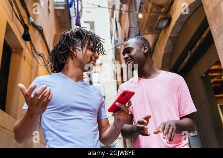 attraente giovane uomo latino-americano a piedi strade con un amico africano. Stanno parlando e sorridendo mentre controllano uno smartphone rosso Foto Stock
