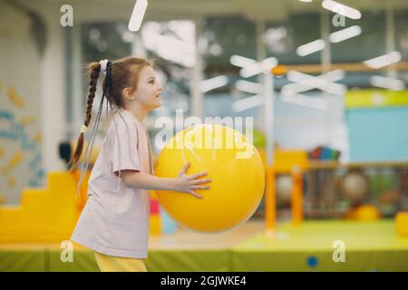 Bambina che gioca e fa le esercitazioni con la sfera gialla grande nella palestra all'asilo o alla scuola elementare. Concetto di sport e fitness per bambini Foto Stock