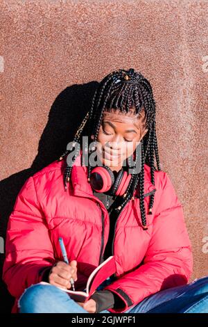 Bella donna africana che indossa giacca rossa con cuffie all'aperto. Donna nera che scrive in un taccuino Foto Stock