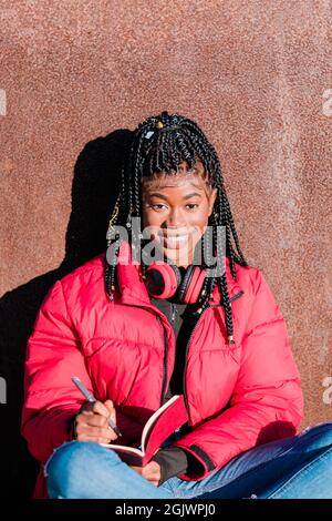 Bella donna africana che indossa giacca rossa con cuffie all'aperto. Donna nera che scrive in un notebook guardando la fotocamera Foto Stock