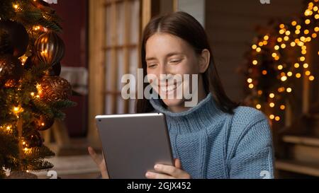 Sorridendo giovane donna caucasica usando la tavoletta seduta vicino all'albero di Natale. Foto Stock