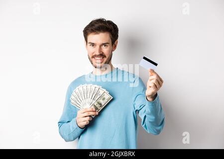 Bel giovane uomo sorridente e che offre il pagamento in contanti e senza contatto, mostrando denaro con carta di credito in plastica, in piedi su sfondo bianco Foto Stock