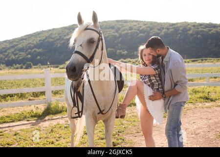 Il giovane uomo barbuto aiuta una giovane donna a salire su un cavallo bianco. L'uomo e la donna trascorrono del tempo insieme nel ranch Foto Stock
