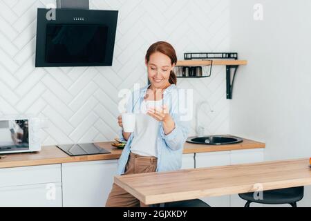 Giovane donna attraente in cucina con una tazza di caffè comunica messaggi al telefono. Foto Stock