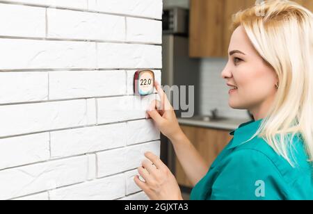 donna che regola la temperatura di riscaldamento con un moderno termostato wireless installato sulla parete bianca a casa. Concetto intelligente di regolazione del riscaldamento domestico Foto Stock