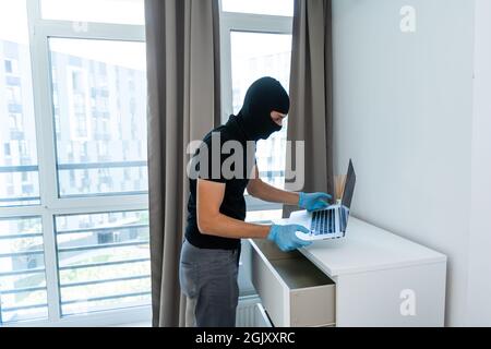 Ladro con balaclava nera rubare laptop. Il burglar commette un crimine in appartamento di lusso Foto Stock