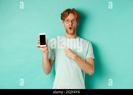 Dai un'occhiata. Bel ragazzo rosso in occhiali che punta il dito sullo schermo vuoto dello smartphone, mostrando la promozione online, in piedi stupito sopra il turchese Foto Stock