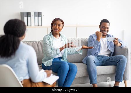 Annoiato donna nera lamentarsi di un uso eccessivo dello smartphone del suo ragazzo durante la consultazione psicologica in clinica Foto Stock