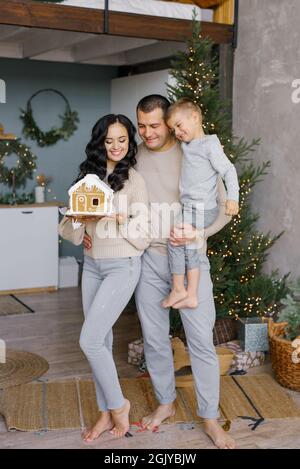 Una giovane famiglia felice con il figlio sta in piedi nella loro cucina, decorata per Natale, e ammirando una casa di pan di zenzero preparata con il proprio ha Foto Stock