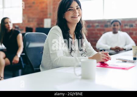 Donna d'affari creativa sorridente mentre si siede in ufficio con i suoi colleghi in background. Felice giovane donna d'affari che guarda la macchina fotografica mentre Foto Stock