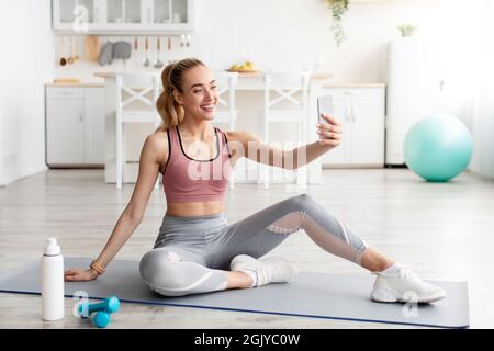 Amichevole sorridente caucasico giovane bionda fitness blogger guardare il telefono cellulare webcam e fare flusso live di allenamento Foto Stock