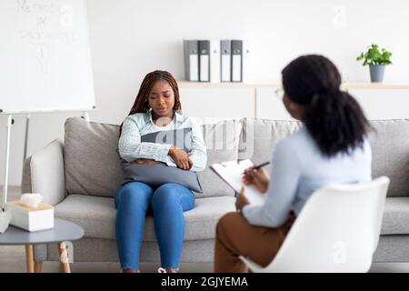 Donna nera depressa seduta sul divano, putting cuscino, avendo consultato lo psicologo alla clinica moderna Foto Stock