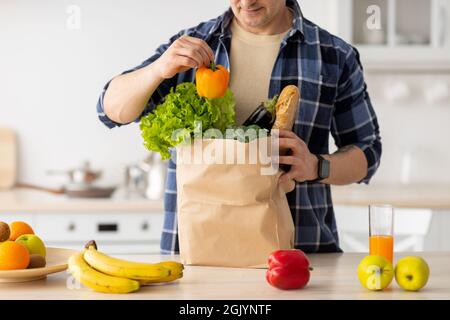 Uomo maturo disimballaggio sacchetto di carta con verdure fresche e frutta dopo la spesa, in piedi in cucina, raccolto Foto Stock