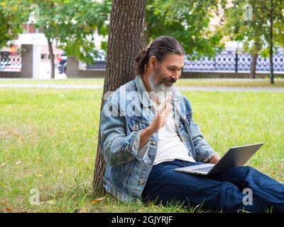 Foto di un uomo di mezza età durante una videochiamata online. È seduto sull'erba nel parco cittadino con un computer portatile. Agitava la mano - Ciao. Foto Stock