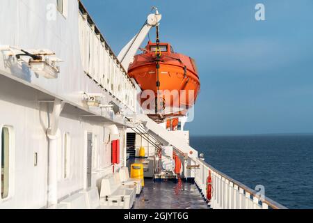 Orange vita barca appesa su una gru su un ponte di barca a vela oceano con orizzonte oceano nella parte posteriore e ringhiera di legno di fronte. Foto Stock