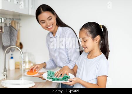 Mano d'aiuto. Ragazza asiatica graziosa che aiuta la madre in cucina, lavando e strofinando i piatti insieme, spazio di copia Foto Stock