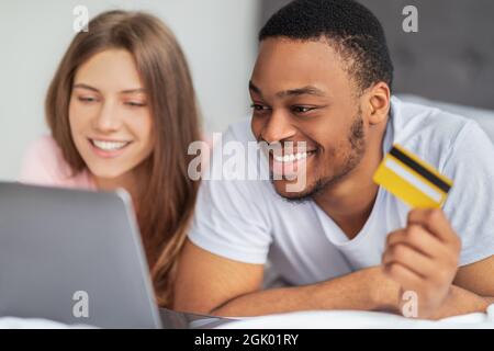 Ritratto di felice coppia multirazziale con carta di credito e computer portatile sdraiato a letto, shopping online da casa Foto Stock