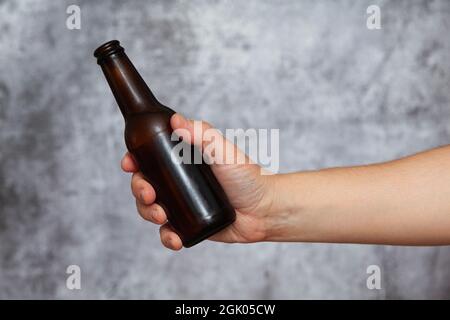 Mano destra di un uomo che tiene una bottiglia senza etichetta di birra fresca con gocce di condensa su uno sfondo grigio testurizzato. Foto Stock