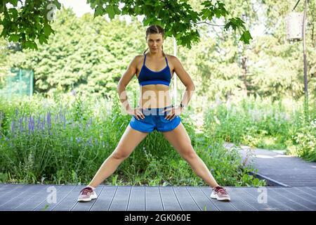 Una ragazza sportiva in una tuta blu si allena nel parco. Si alza in posa con le gambe larghe. Foto Stock