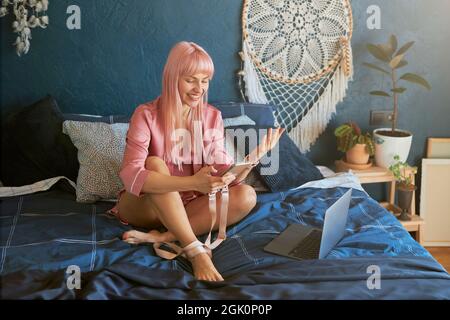 Signora gioiosa con i capelli rosa in abito di seta tiene il telefono seduto vicino a un computer portatile su un grande letto Foto Stock