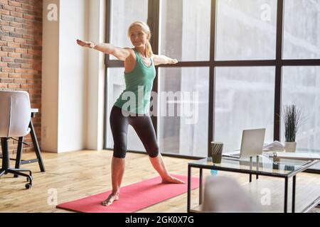 Scatto a tutta lunghezza di donna matura sportiva in piedi in posizione Warrior mentre pratica yoga a casa in appartamento moderno Foto Stock