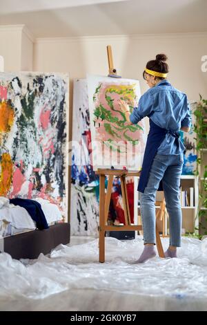 Ripresa a tutta lunghezza di artista femminile di talento in grembiule pittura su tela in casa studio laboratorio Foto Stock