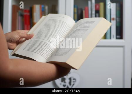 Una mano di donna che legge un libro vicino ad una libreria Foto Stock