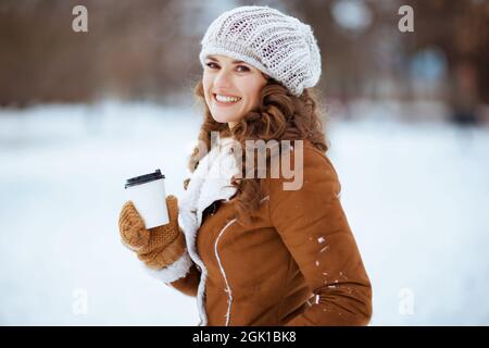 Ritratto di felice elegante donna di mezza età con guanti e tazza di cioccolata calda in un cappello a maglia e cappotto di pelle di pecora all'aperto nel parco della città in inverno. Foto Stock