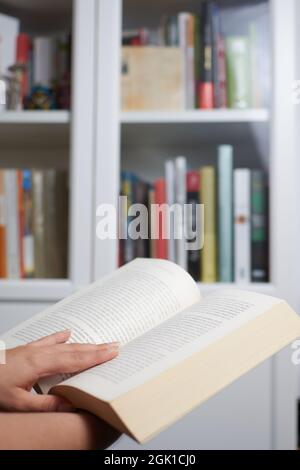 Una mano di donna che legge un libro vicino ad una libreria Foto Stock